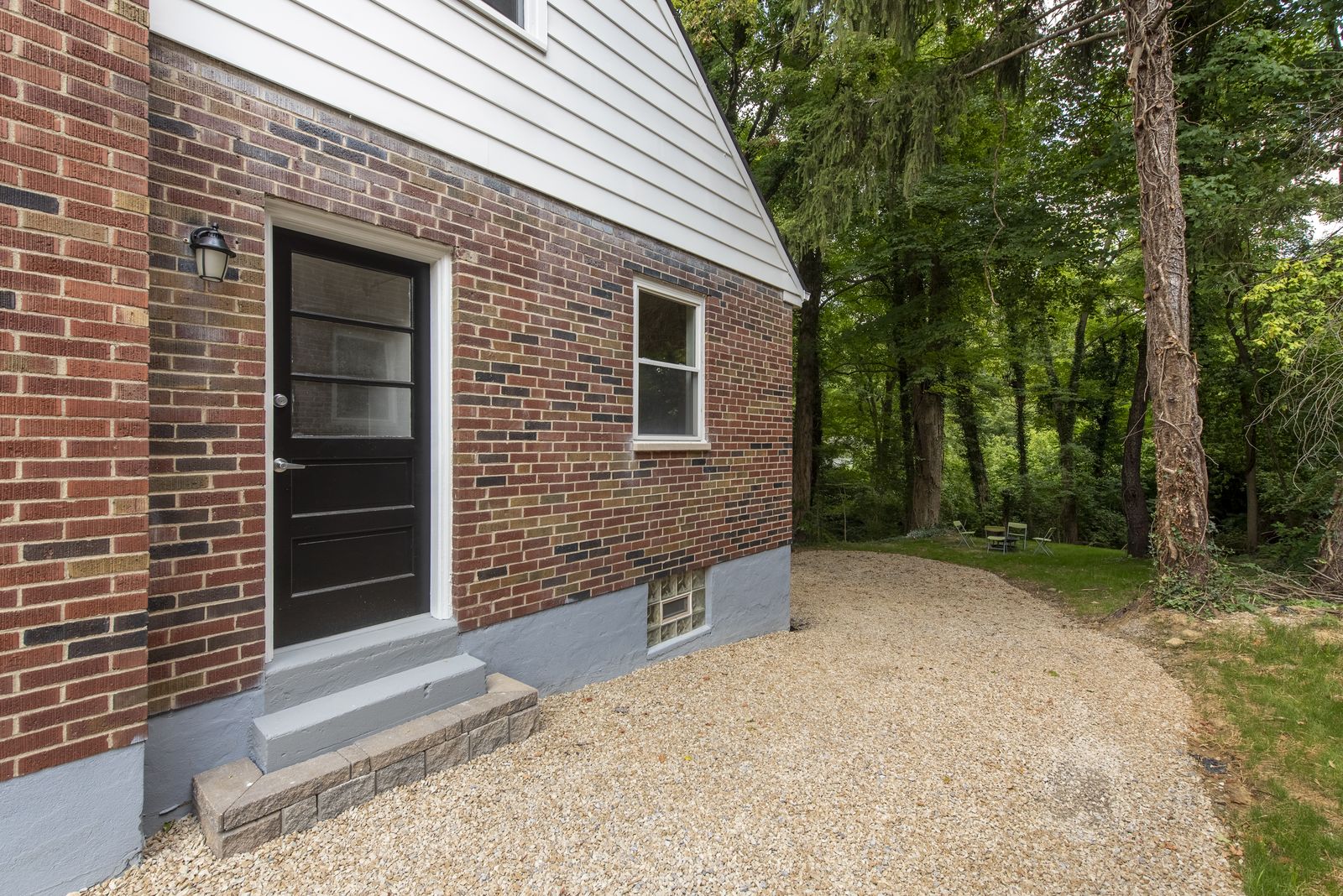 Renovated side entry of a brick Cape Cod home with a black paneled door, white siding, and a new gravel walkway