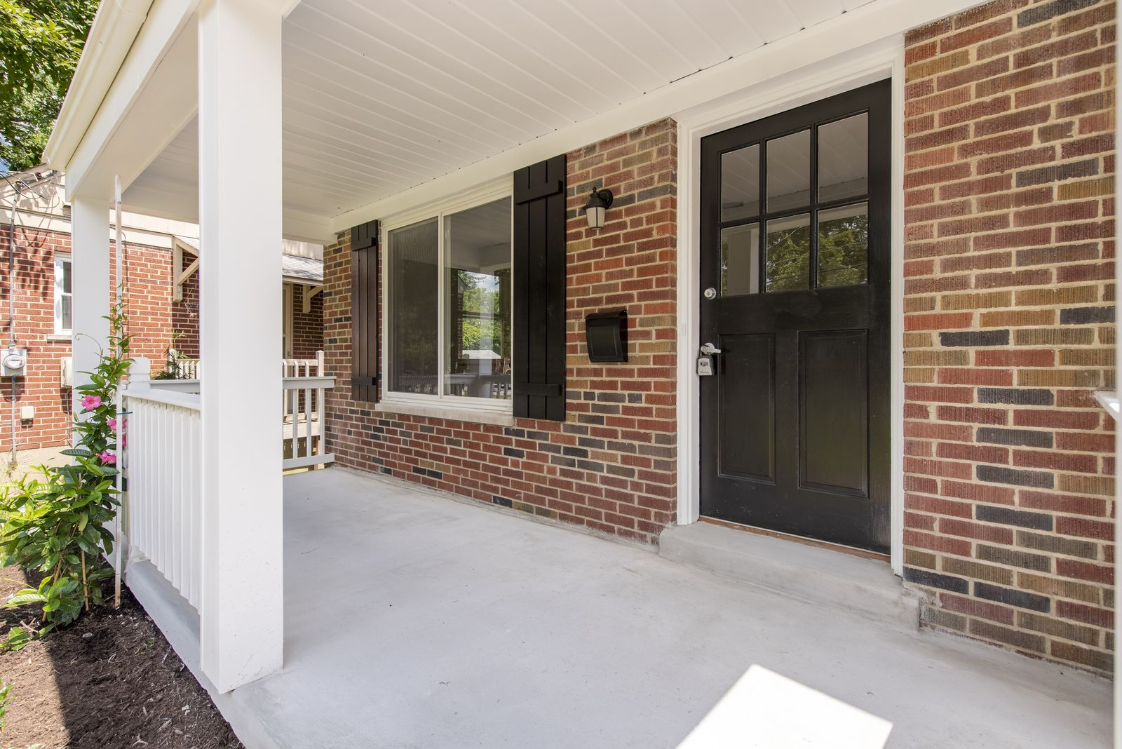 Covered front porch with white columns and railing, brick facade, black front door, and matching black shutters