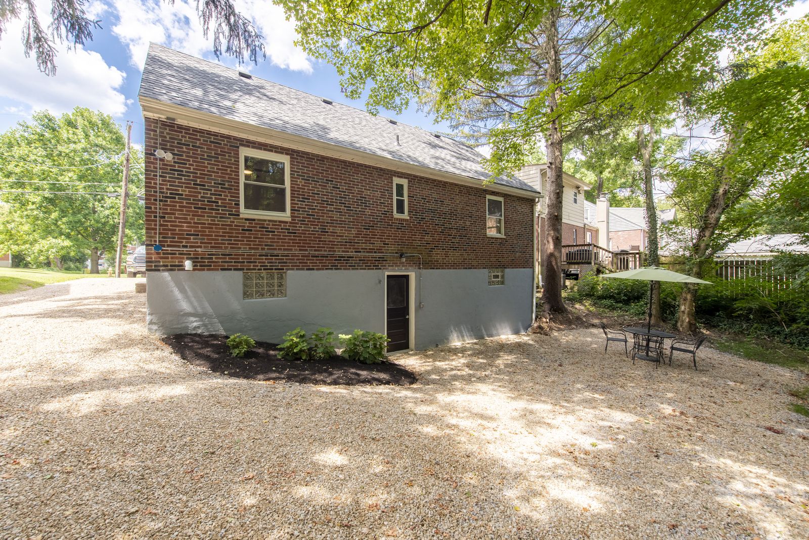 Rear brick exterior of the renovated home with a painted foundation, walk-out basement door, gravel patio, and mature trees