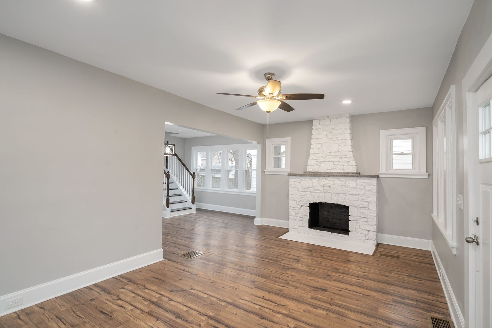 Bright living room with a white-painted stone fireplace, new wood-look flooring, and a refinished staircase in the background