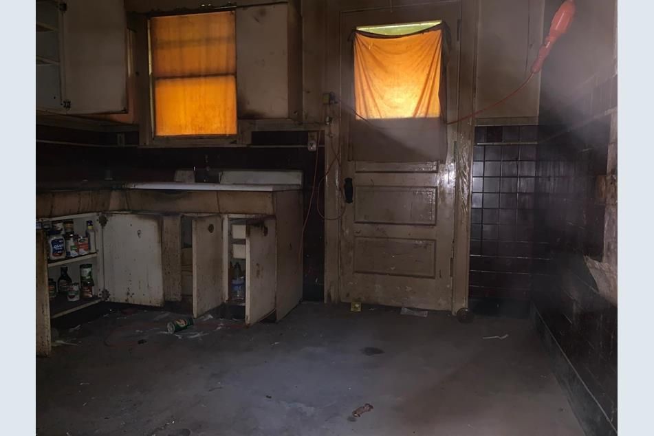 Kitchen before renovation, with grimy old cabinets, covered windows, and worn flooring
