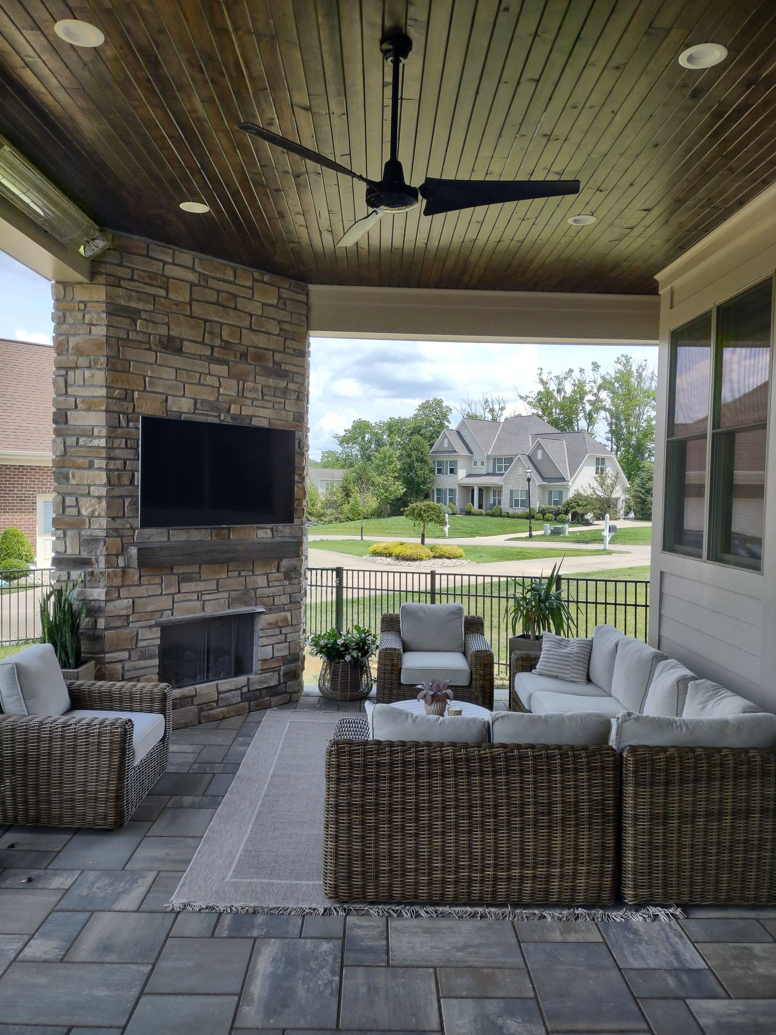 Covered porch lounge with stone fireplace, wall-mounted TV and wicker seating under a stained wood ceiling