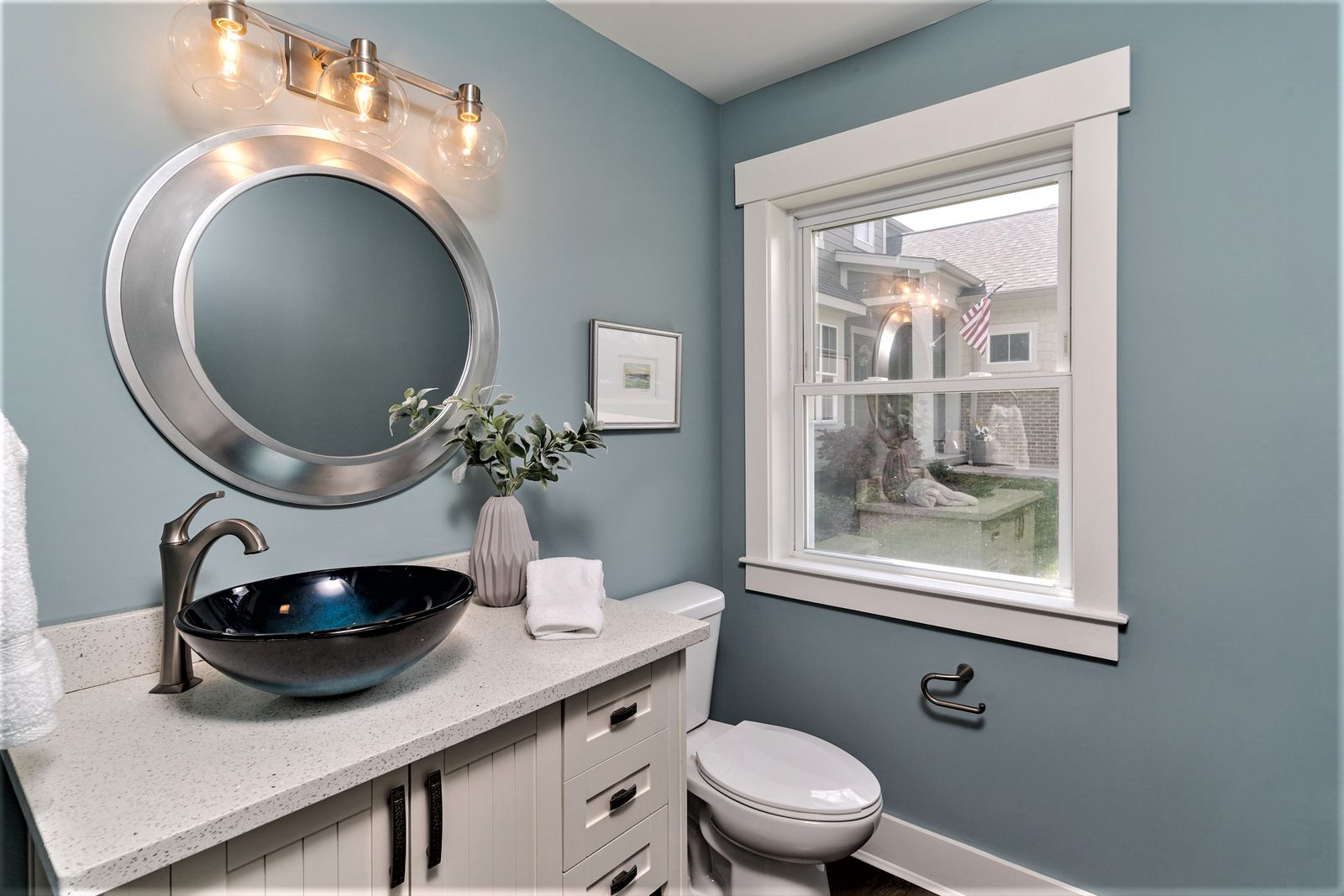 Powder room with blue walls, dark vessel sink, round mirror, and white vanity