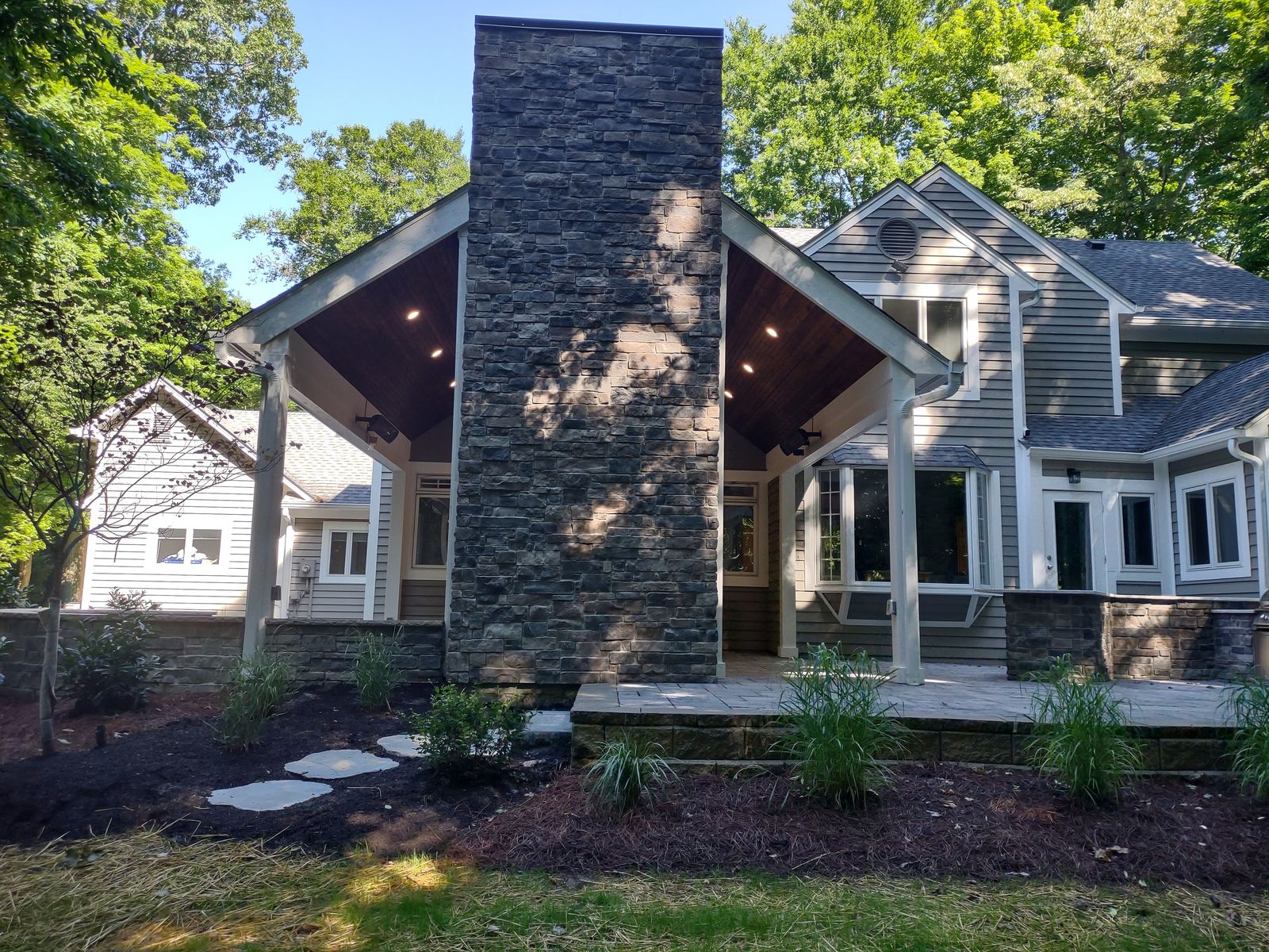 Covered porch with tall stone chimney and vaulted wood ceiling behind home